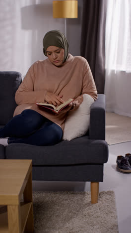 Video-Vertical-De-Una-Mujer-Musulmana-Usando-Hijab-Sentada-En-Un-Sofá-En-Casa-Leyendo-O-Estudiando-El-Corán